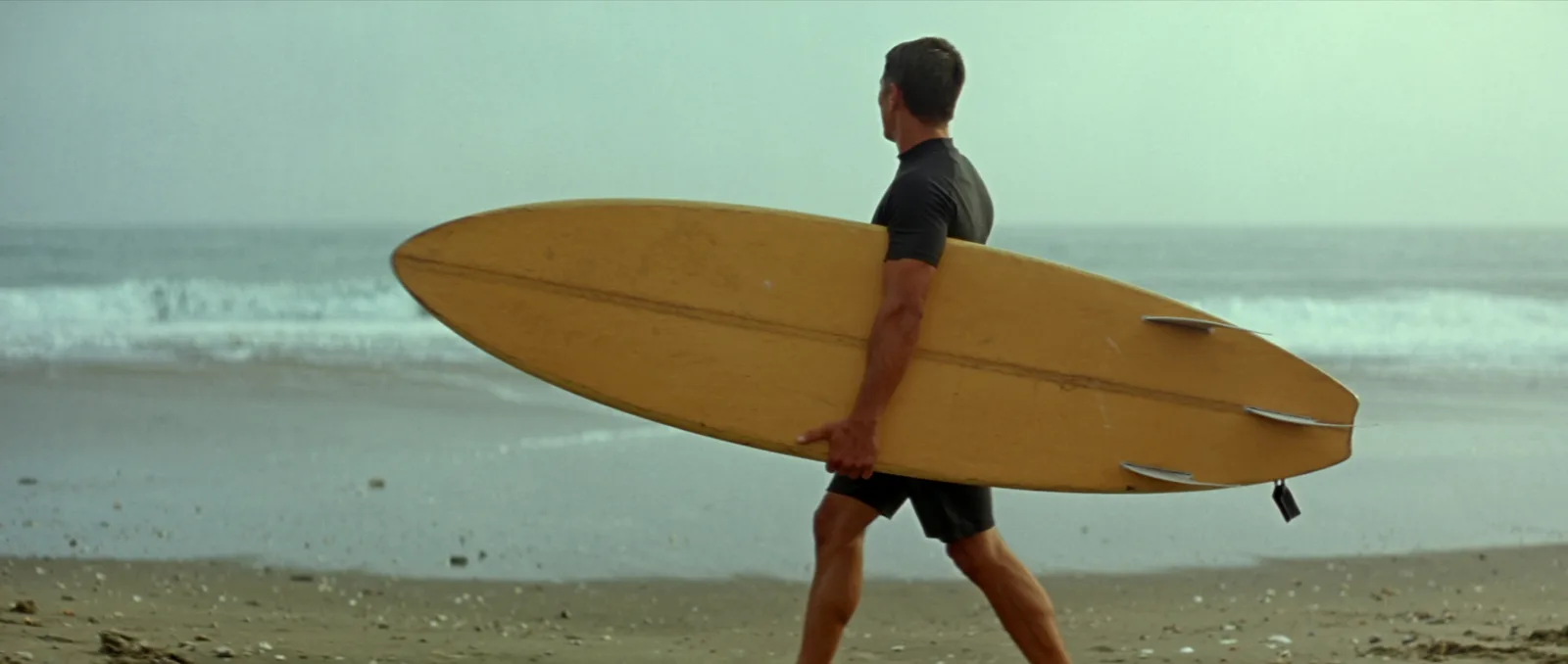 Surfboard weight — a surfer carrying a longboard under one arm across wet sand