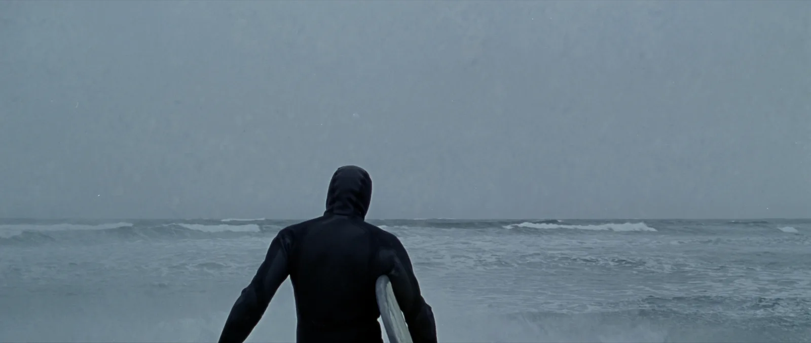 Wetsuit thickness guide — a surfer in a thick hooded winter wetsuit wading into cold grey water
